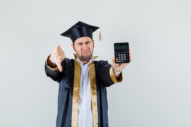 Młody mężczyzna trzyma kalkulator, pokazując kciuk w dół w mundurze absolwenta i wyglądający na rozczarowanego. przedni widok.