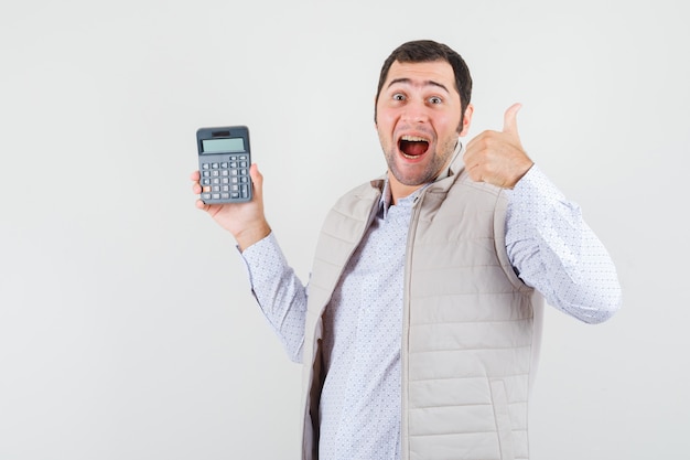 Młody mężczyzna trzyma kalkulator w jednej ręce, pokazując kciuk w beżowej kurtce i czapce i wygląda optymistycznie. przedni widok.