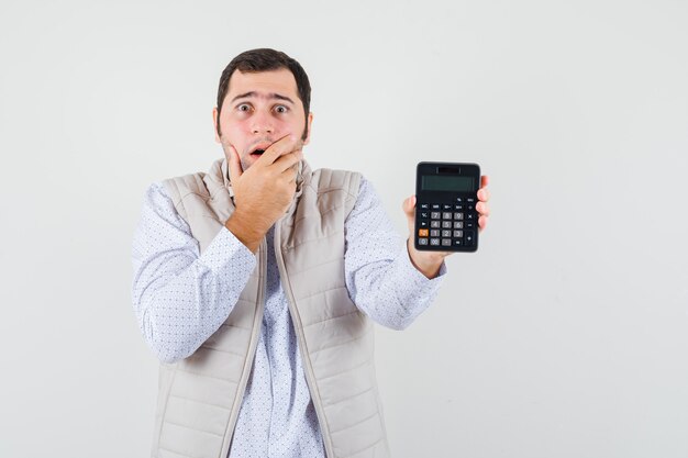 Młody mężczyzna trzyma kalkulator w jednej ręce, zakrywając usta ręką w beżowej kurtce i czapce i wygląda na zaskoczonego. przedni widok.