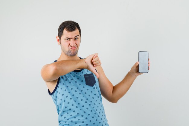 Młody mężczyzna trzyma telefon komórkowy, pokazując kciuk w dół w niebieskim podkoszulku i patrząc niezadowolony