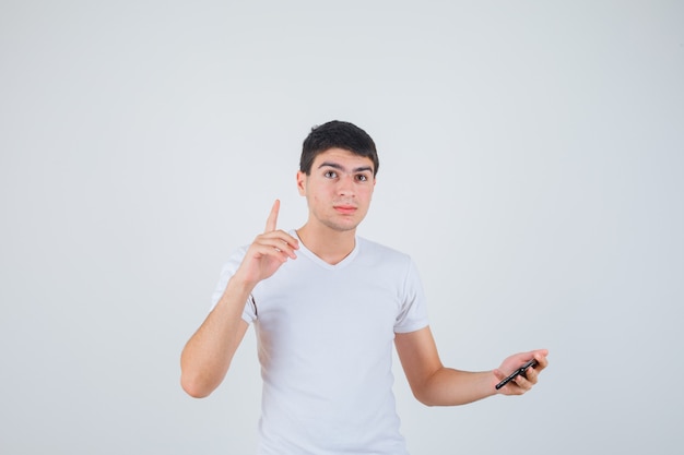 Młody mężczyzna trzyma telefon, wskazując w t-shirt i patrząc pewny siebie, przedni widok.