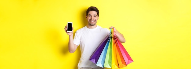 Młody mężczyzna trzyma torby na zakupy i pokazuje aplikację pieniężną na ekranie telefonu komórkowego stojącą nad krzykiem