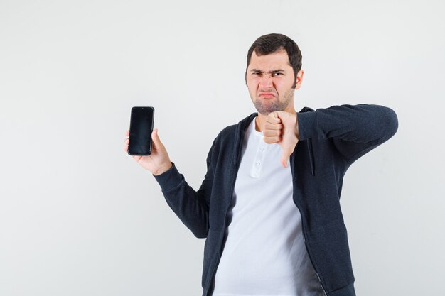Młody mężczyzna trzymający smartfon, pokazujący kciuk w dół w białej koszulce i czarnej bluzie z zamkiem błyskawicznym, wyglądający na niezadowolonego, widok z przodu.
