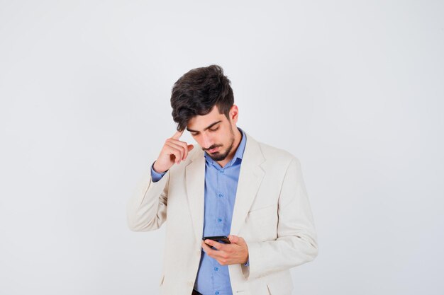 Młody mężczyzna trzymający smartfona i kładący palec wskazujący na uchu w niebieskiej koszulce i białej marynarce i patrzący poważnie