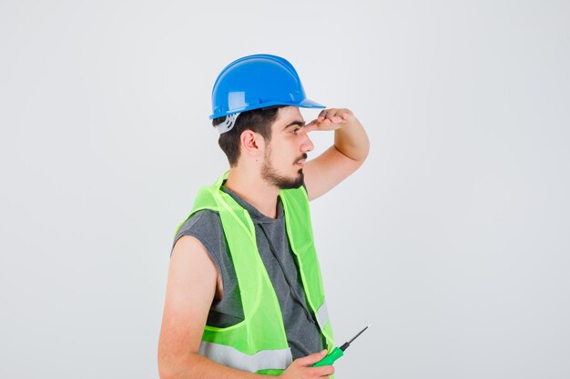 Młody mężczyzna trzymający śrubokręt i odwracający wzrok w mundurze budowlanym i patrzący poważnie