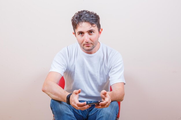 Młody mężczyzna trzymający telefon komórkowy siedząc na krześle w koszulce, dżinsach