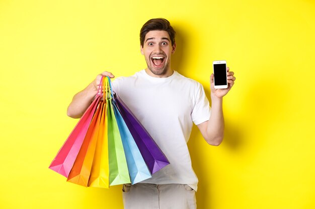 Młody mężczyzna trzymający torby na zakupy i pokazujący ekran telefonu komórkowego, aplikację pieniędzy, stojący na żółtym tle