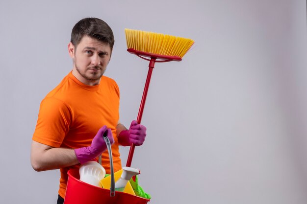 Młody mężczyzna ubrany w pomarańczową koszulkę i gumowe rękawiczki trzymający wiadro z narzędziami do czyszczenia i mopem, wyglądający na pewnego siebie gotowego do clen, stojącego nad białą ścianą