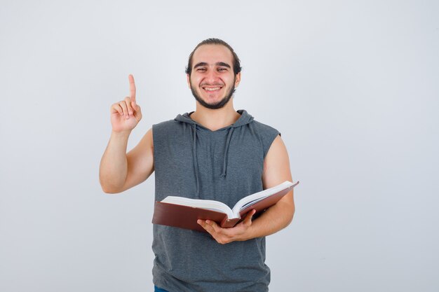 Młody mężczyzna w bez rękawów z kapturem, trzymając książkę, wskazując w górę i patrząc szczęśliwy, widok z przodu.