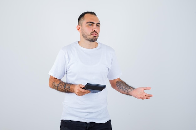 Młody mężczyzna w białej koszulce i czarnych spodniach, trzymający kalkulator i wyciągający rękę w pytający sposób i wyglądający poważnie