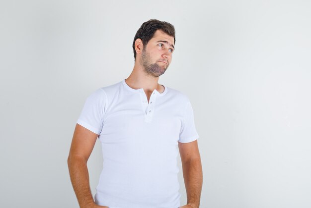 Młody mężczyzna w białej koszulce odwracający wzrok z rękami w kieszeniach i wyglądający na beznadziejnego
