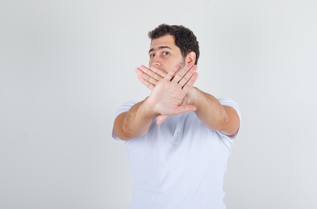 Młody mężczyzna w białej koszulce pokazuje gest stop i wygląda na zniesmaczonego