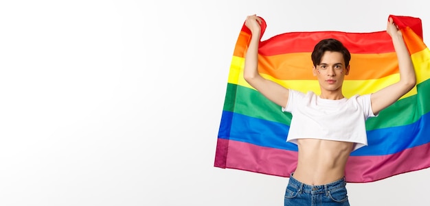 Młody mężczyzna w crop top z brokatem na twarzy podnoszący flagę dumy z pewnymi emocjami queer person sm