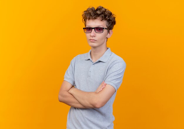 Młody mężczyzna w czarnych okularach na sobie szarą koszulkę polo z poważną twarzą ze skrzyżowanymi rękami na piersi, stojąc na pomarańczowej ścianie