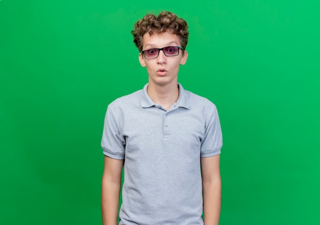 Młody mężczyzna w czarnych okularach ubrany w szarą koszulkę polo zaskoczony i zdumiony stojąc nad zieloną ścianą