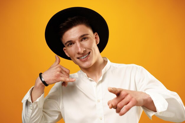 Młody mężczyzna w czarnym kapeluszu pokazujący znak „Zadzwoń do mnie” przed żółtym backgorund