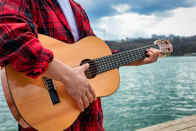 Młody mężczyzna w czerwonej koszuli gra na gitarze w pobliżu jeziora w parku