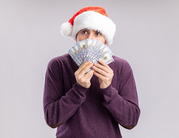 Młody mężczyzna w fioletowym swetrze i santa hat trzymając gotówkę zakrywającą twarz pieniędzmi patrząc na bok zmartwiony stojąc na białym tle