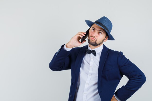 Młody mężczyzna w garniturze, kapeluszu rozmawia przez telefon komórkowy i patrząc niezdecydowanie, widok z przodu.
