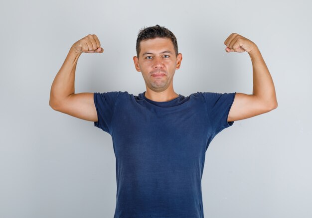 Młody mężczyzna w granatowej koszulce pokazujący mięśnie i silnie wyglądający, widok z przodu.