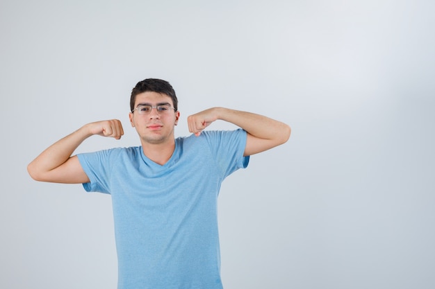 Młody mężczyzna w koszulce pokazując gest mięśni i patrząc pewnie, widok z przodu.
