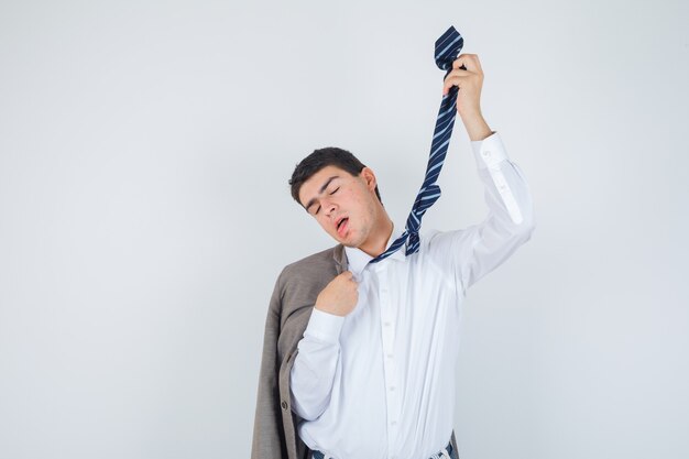 Młody mężczyzna w koszuli, kurtce, krawacie w paski wykonujący gest samobójczy z krawatem i wyglądający beznadziejnie