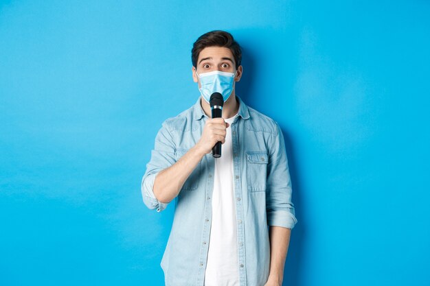Młody mężczyzna w masce medycznej, przemawiający, trzymający mikrofon i patrzący zdezorientowany, stojący na niebieskim tle.