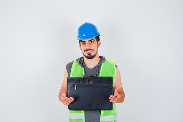 Młody mężczyzna w mundurze budowlanym trzymający walizkę na narzędzia budowlane obiema rękami i wyglądający na szczęśliwego