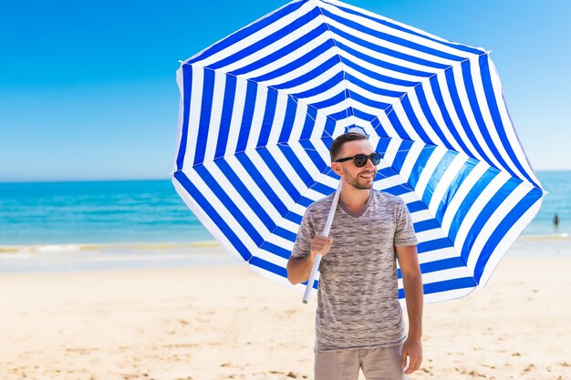 Młody mężczyzna w okularach przeciwsłonecznych spacerujący po plaży ze słonecznym parasolem słonecznym