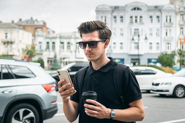 Młody mężczyzna w okularach przeciwsłonecznych ze smartfonem i kawą w mieście