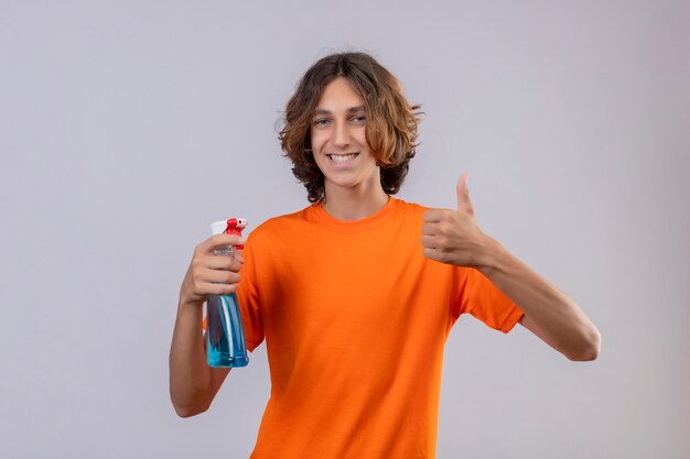 Młody mężczyzna w pomarańczowej koszulce gospodarstwa spray do czyszczenia, uśmiechając się radośnie patrząc na kamery pokazując kciuki stojąc na białym tle