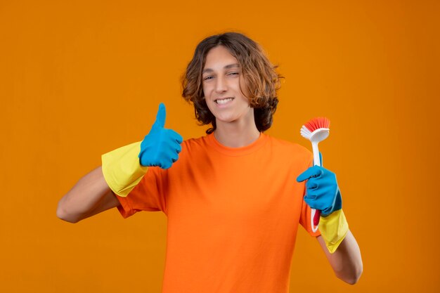 Młody mężczyzna w pomarańczowej koszulce w gumowych rękawiczkach, trzymając pędzel do szorowania uśmiechnięty z radosną buźką pokazując kciuki do góry stojąc na żółtym tle