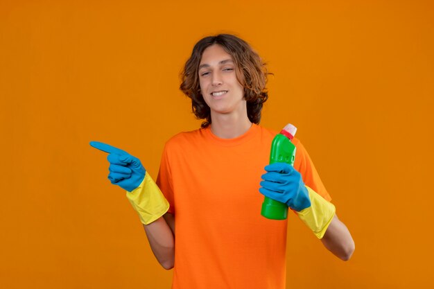 Młody mężczyzna w pomarańczowej koszulce w gumowych rękawiczkach, trzymając szczotkę do szorowania, wskazując w bok, patrząc pewnie uśmiechnięty stojący na żółtym tle