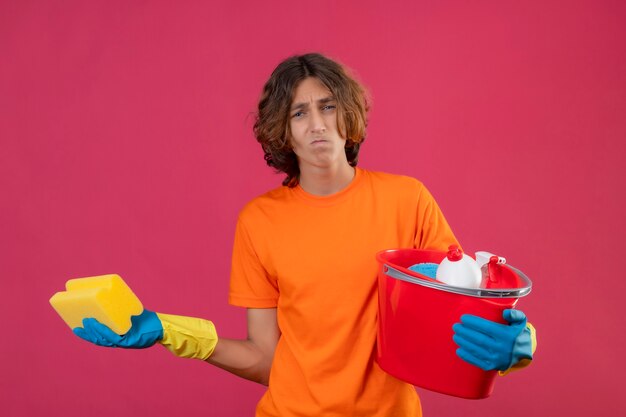 Młody mężczyzna w pomarańczowej koszulce w gumowych rękawiczkach trzymający gąbkę i wiadro z narzędziami do czyszczenia wzruszający ramionami, wyglądający na niepewnego i zdezorientowanego, bez odpowiedzi, rozkładający dłonie na stojąco