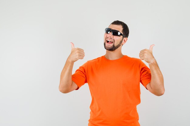 Młody mężczyzna w pomarańczowym t-shirt pokazano podwójne kciuki w górę i patrząc wesoły, widok z przodu.