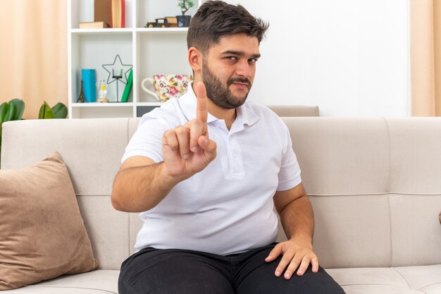 Młody mężczyzna w swobodnym ubraniu z pewnym siebie wyrazem twarzy pokazującym gest ostrzegawczy palcem wskazującym, siedzący na kanapie w jasnym salonie