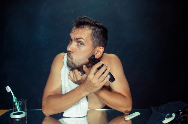 Młody mężczyzna w sypialni, siedzący przed lustrem, drapiąc się w brodę w domu. Koncepcja ludzkich emocji