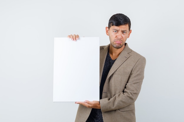 Młody mężczyzna w szaro-brązowej kurtce stojącej z trzymając czysty papier i patrząc smutno, widok z przodu.