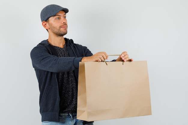 Bezpłatne zdjęcie młody mężczyzna w t-shirt, marynarce, dżinsach, czapce dostarczającej papierową torbę, widok z przodu.