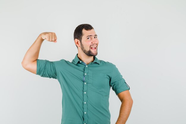 Młody mężczyzna w zielonej koszuli pokazuje mięśnie ramion i wygląda na szczęśliwego, widok z przodu.