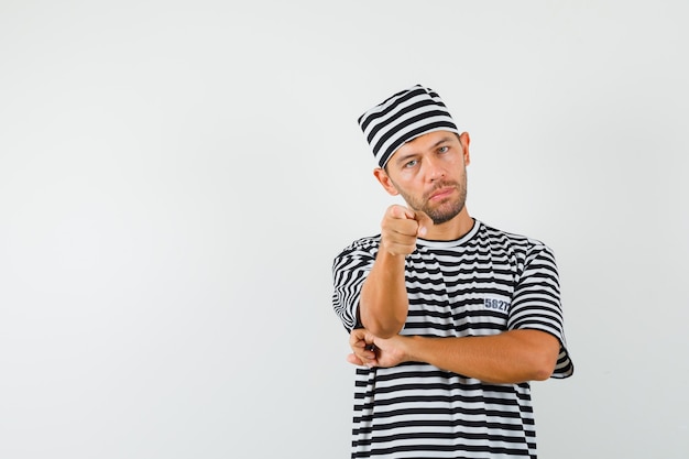 Bezpłatne zdjęcie młody mężczyzna, wskazując na aparat w kapelusz w paski t-shirt i patrząc pewnie