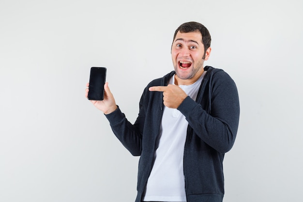 Młody mężczyzna wskazujący smartfon, uśmiechnięty w białej koszulce i czarnej bluzie z zamkiem błyskawicznym i wyglądający na szczęśliwego. przedni widok.
