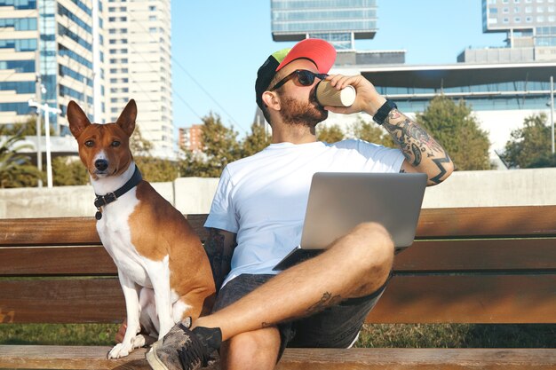 Młody mężczyzna z brodą i tatuażami i laptopem na kolanach pije kawę z papierowego kubka, a obok niego siedzi pies