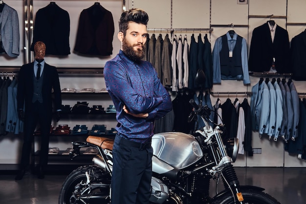 Młody modny mężczyzna z brodą modny ubrany pozowanie w pobliżu motocykla sportowego retro w sklepie z odzieżą męską.