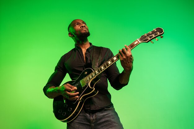 Młody muzyk afro-amerykański gra na gitarze jak gwiazda rocka na gradientowym tle zielono-żółtym. Koncepcja muzyki, hobby, festiwalu, pleneru. Radosny facet improwizujący, śpiewający piosenkę.