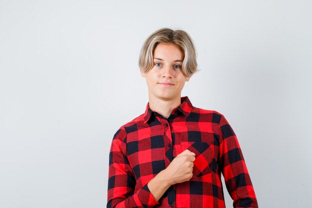 Młody nastolatek chłopiec trzymając pięść na klatce piersiowej w kraciastej koszuli i wyglądający pewnie, widok z przodu.