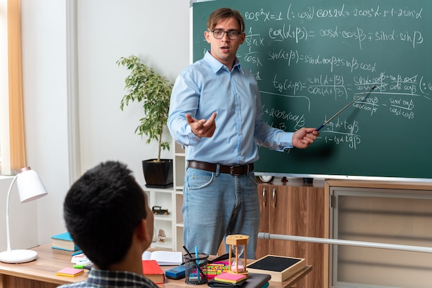 Młody nauczyciel płci męskiej w okularach wyglądający na zdezorientowanego i niezadowolonego wyjaśniającego lekcję uczniom, stojący w pobliżu tablicy z formułami matematycznymi w klasie