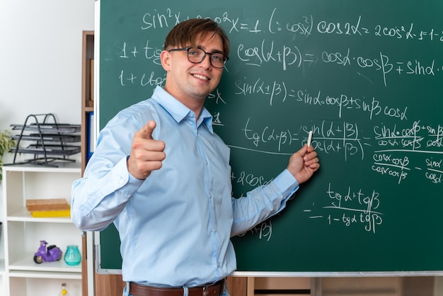 Młody nauczyciel płci męskiej w okularach z kredą wyjaśniającą lekcję szczęśliwą i pozytywnie uśmiechniętą stojącą w pobliżu tablicy z formułami matematycznymi w klasie