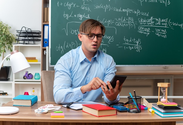 Młody nauczyciel w okularach, piszący wiadomość za pomocą smartfona, wyglądający pewnie siedzący przy szkolnym biurku z książkami i notatkami przed tablicą w klasie