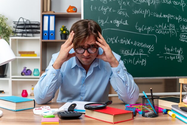Młody nauczyciel w okularach zestresowany i zdenerwowany siedzący przy szkolnej ławce z książkami i notatkami przed tablicą w klasie in